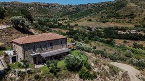 Una casa de piedra con techo de tejas rojas, rodeada de un jardín cuidado y un paisaje ondulado.