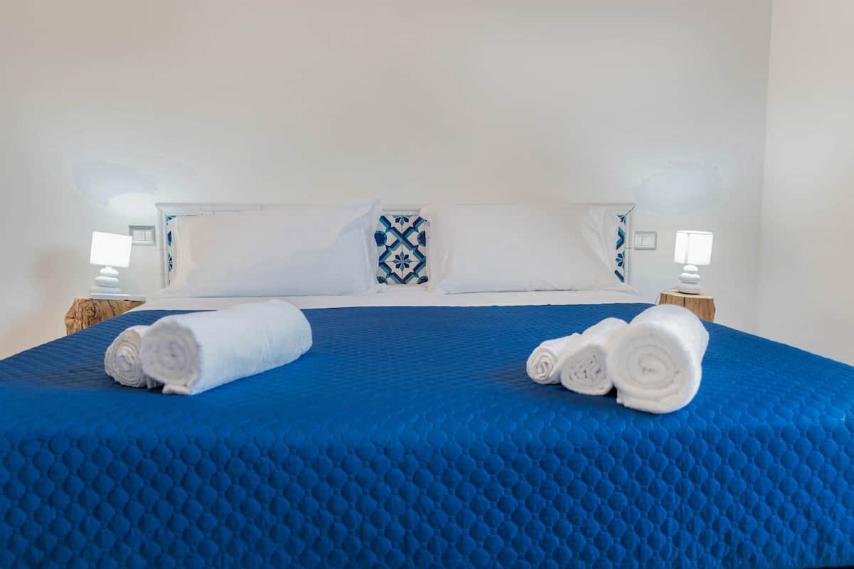 Un lit bien fait, avec une literie bleue et des oreillers blancs, deux tables de chevet avec des lampes, et un coussin à motifs posé sur la tête de lit.