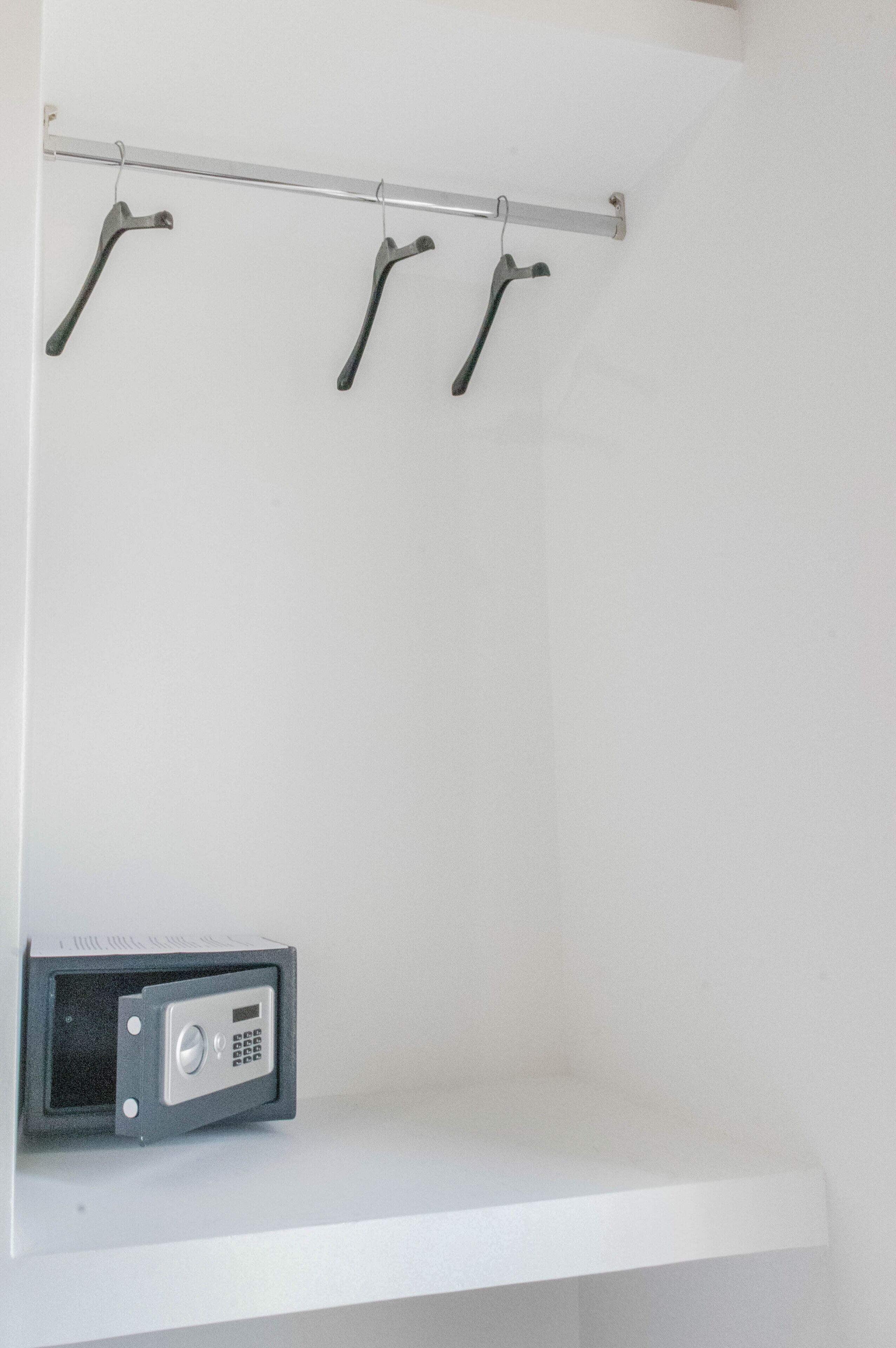 Een witte muur met een metalen rek waarop drie zwarte haken en een klein kluisje met toetsenbord bevestigd zijn.