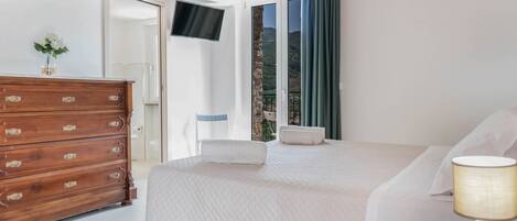 A bedroom with a bed, a wooden dresser, a TV mounted on the wall, and a balcony with a view of the outdoors.