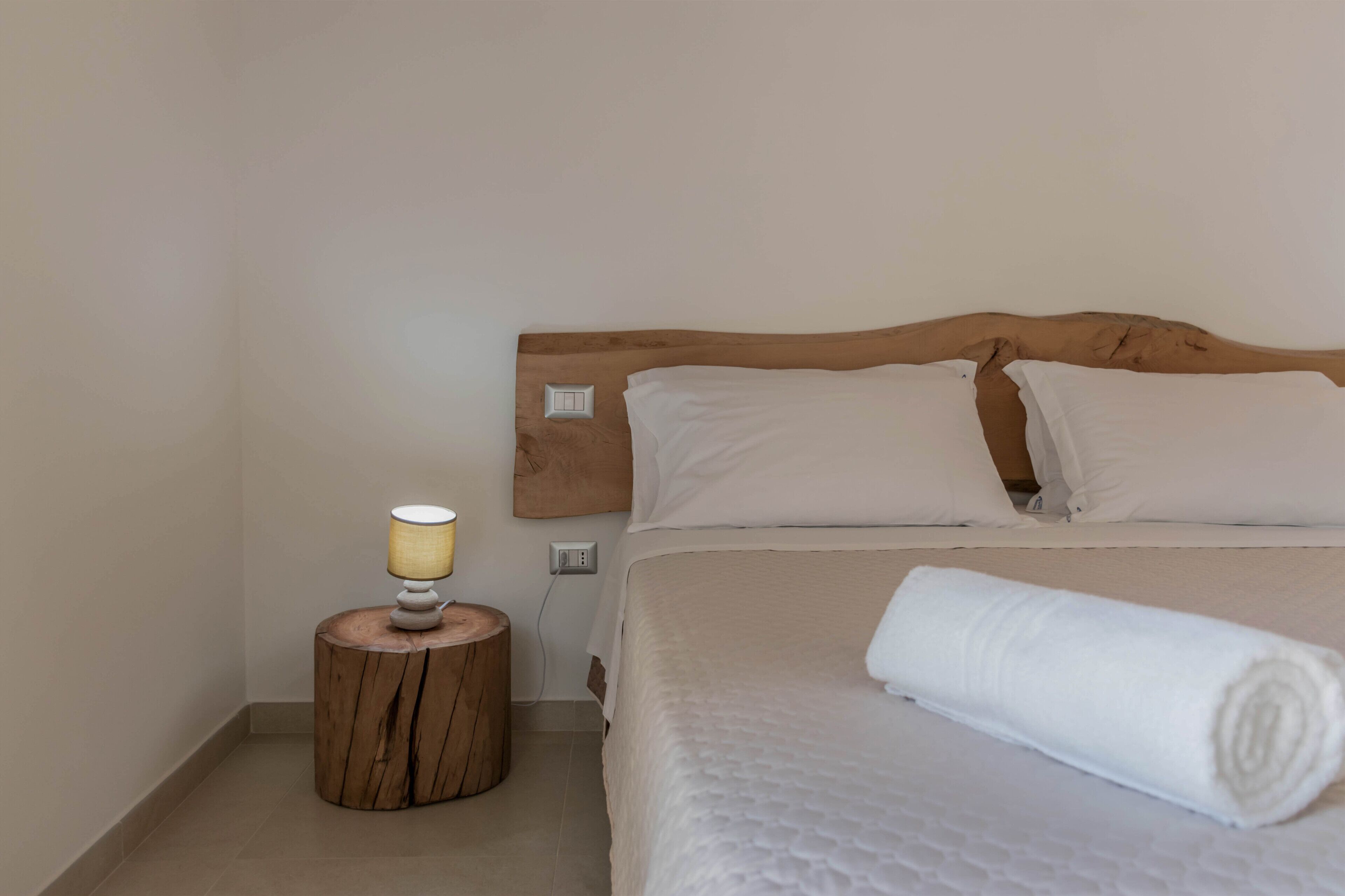 A neatly made bed with a wooden headboard, a bedside lamp on a wooden stool, and a rolled white towel on the bed.