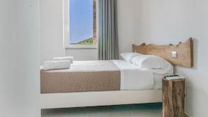 Una cama bien hecha con cabecera de madera, una ventana que ofrece vistas a un paisaje verde y un taburete de madera con un control remoto encima.