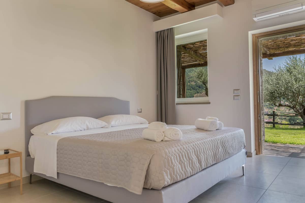 Une chambre moderne avec un grand lit, une tête de lit en bois, une fenêtre donnant sur un espace verdoyant et une porte menant à un espace extérieur.