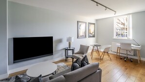 Una sala de estar moderna con un televisor de pantalla plana, un sofá gris, una mesita redonda y un escritorio blanco con una silla.