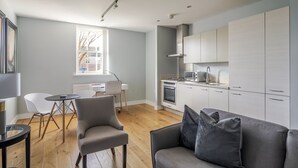 Una sala moderna con un sofá gris, una pequeña mesa de comedor y una silla blanca. Se ve una cocina con gabinetes blancos y una estufa al fondo.