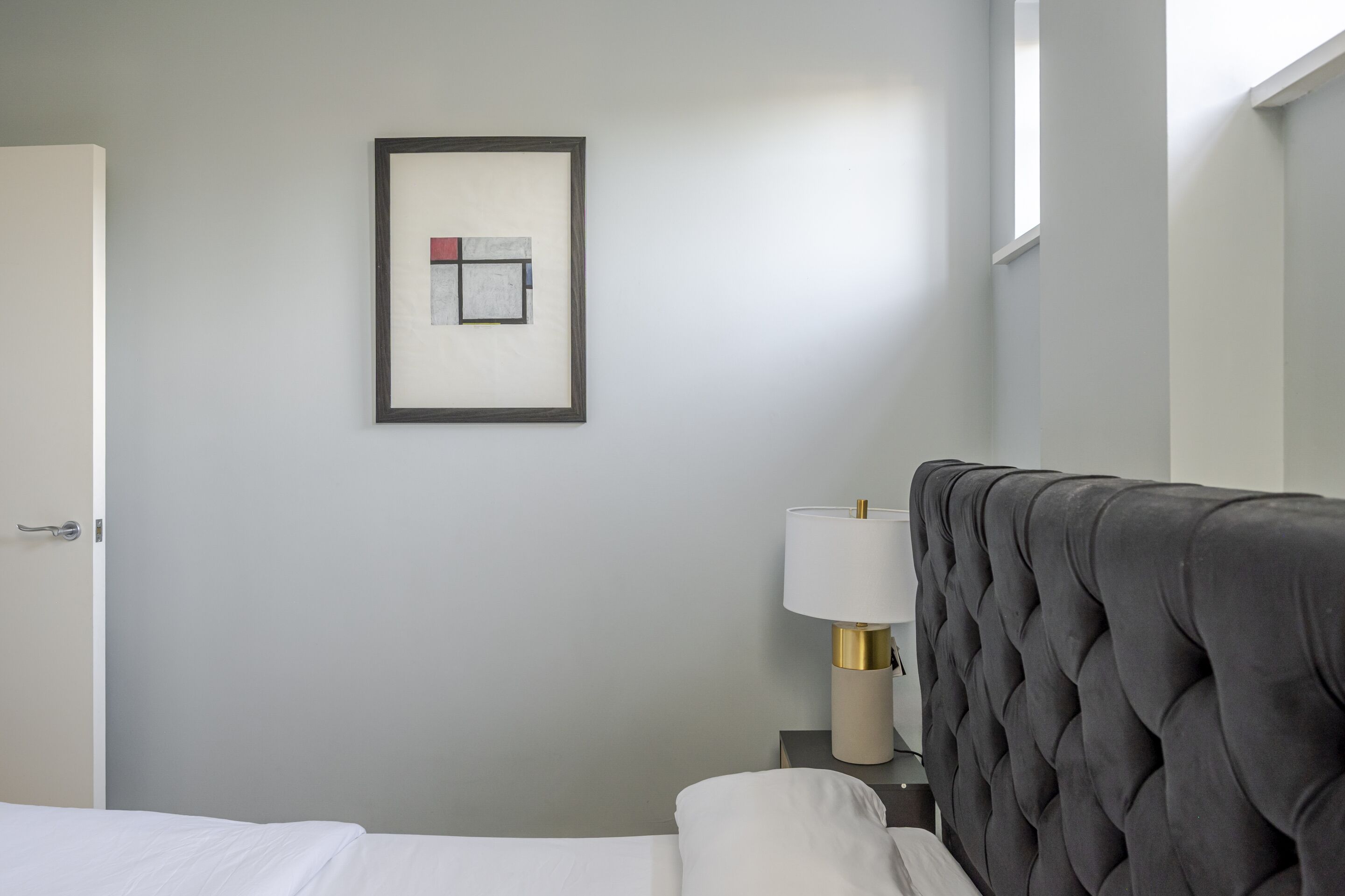 A hotel room with a neatly made bed, a framed artwork on the wall, a bedside lamp, and a window providing natural light.