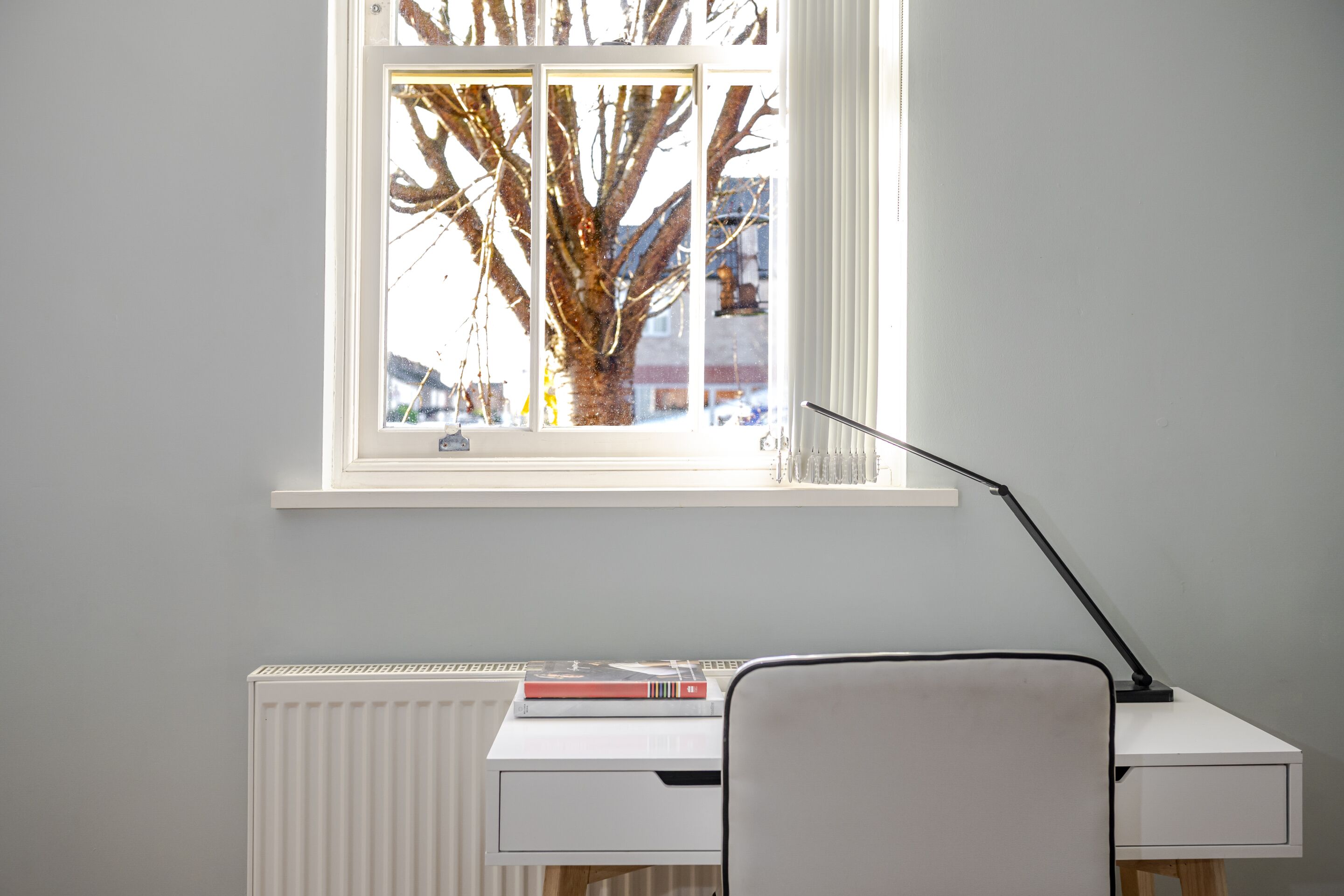 Een kamer met een wit bureau, een radiator, een raam met uitzicht op een boom en gebouwen, en een lamp.