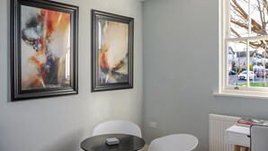 Una habitación con dos cuadros abstractos, un radiador blanco, una mesita con sillas y un ventanal con vistas a coches aparcados y un árbol.