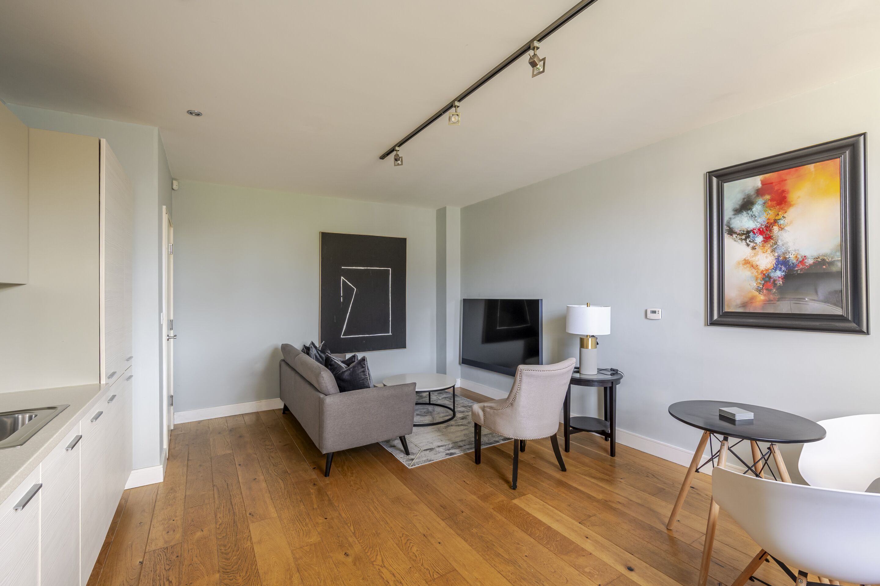 A modern living room with a grey sofa, a small round table, a white armchair, a TV, and a colorful abstract painting on the wall.