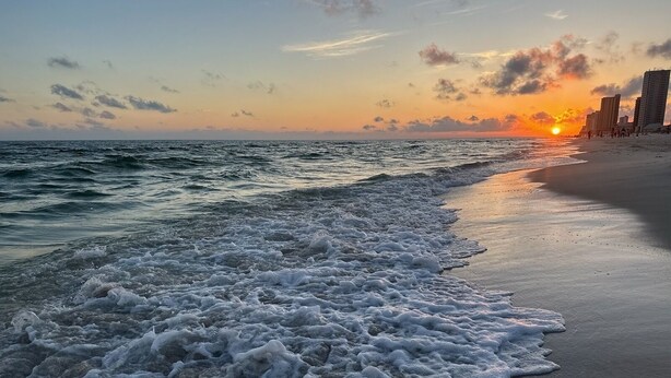 Una playa con olas al atardecer, una orilla arenosa y edificios al fondo.