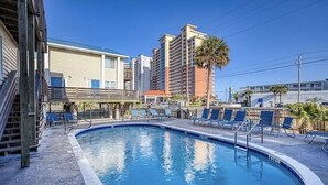 Una piscina con sillas de descanso, una terraza de madera y un edificio de varios pisos al fondo.