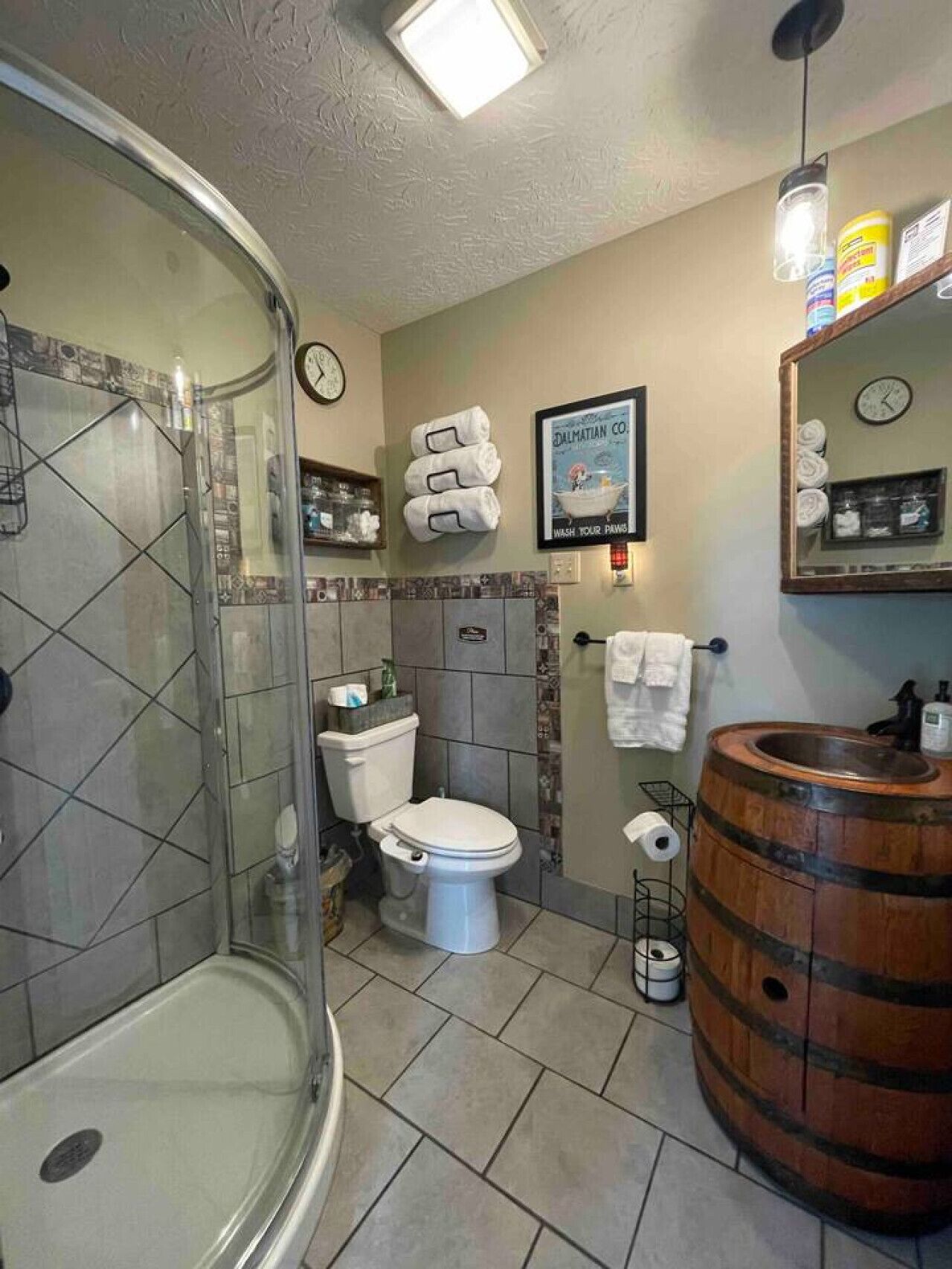 A bathroom with a glass shower enclosure, a toilet, a sink with a wooden barrel, and a mirror with a clock.