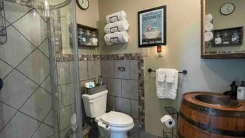 A bathroom with a glass shower enclosure, a toilet, a sink with a wooden barrel, and a mirror with a clock.