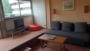 Ein Wohnzimmer mit einer Couch, einem kleinen Holztisch, einem roten Sitzsack, einem Fernseher und einem Fenster mit Jalousien.