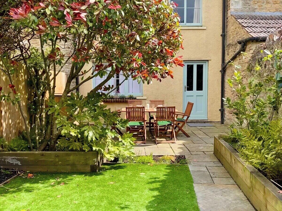 A patio with a table and chairs, surrounded by a garden with a variety of plants and a wooden fence.