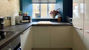 Une cuisine avec un mur d’accent bleu, une fenêtre avec des stores, une horloge et divers appareils électroménagers.