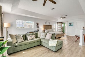 A modern bedroom with a green sectional sofa, a bed with a wooden headboard, a ceiling fan, and a window with blinds. - Tropical Retreat! 2 minutes to Beach and Pool/Jacuzzi! (Panama City Beach)