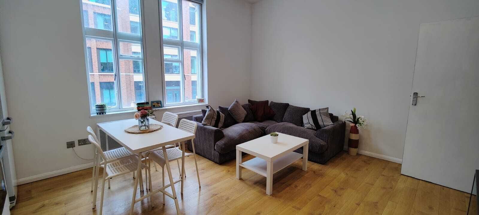 A small, bright living room with a wooden floor, a white table, a striped sofa, and a window with a city view.