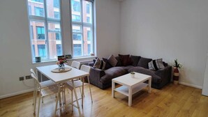 Ein kleines, helles Wohnzimmer mit Holzboden, einem weißen Tisch, einem gestreiften Sofa und einem Fenster mit Blick auf die Stadt.