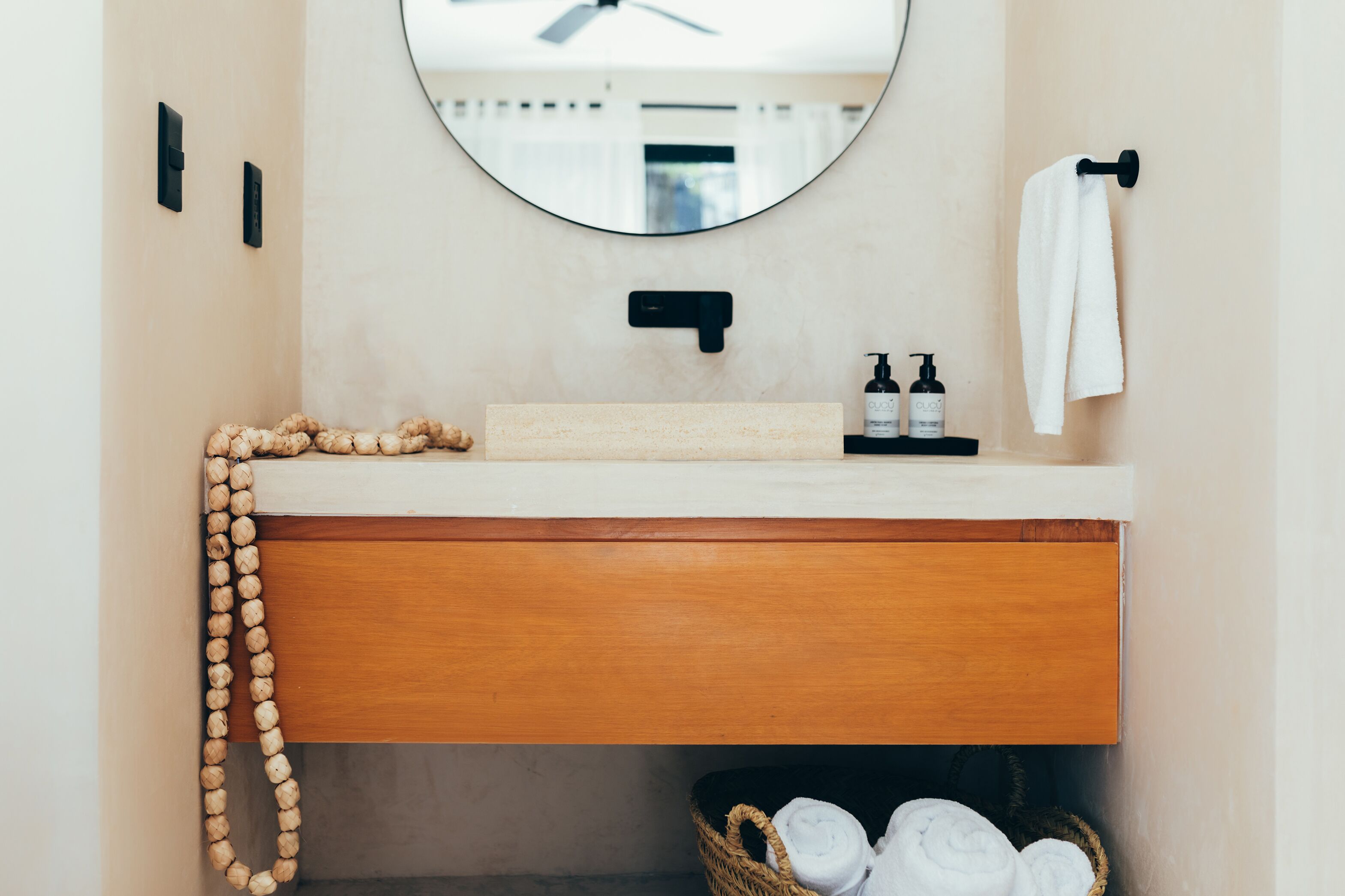 A bathroom with a wooden vanity, a round mirror, a towel hanging on the wall, and a basket of towels on the floor.