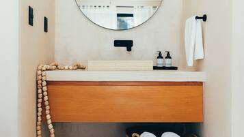 A bathroom with a wooden vanity, a round mirror, a towel hanging on the wall, and a basket of towels on the floor.