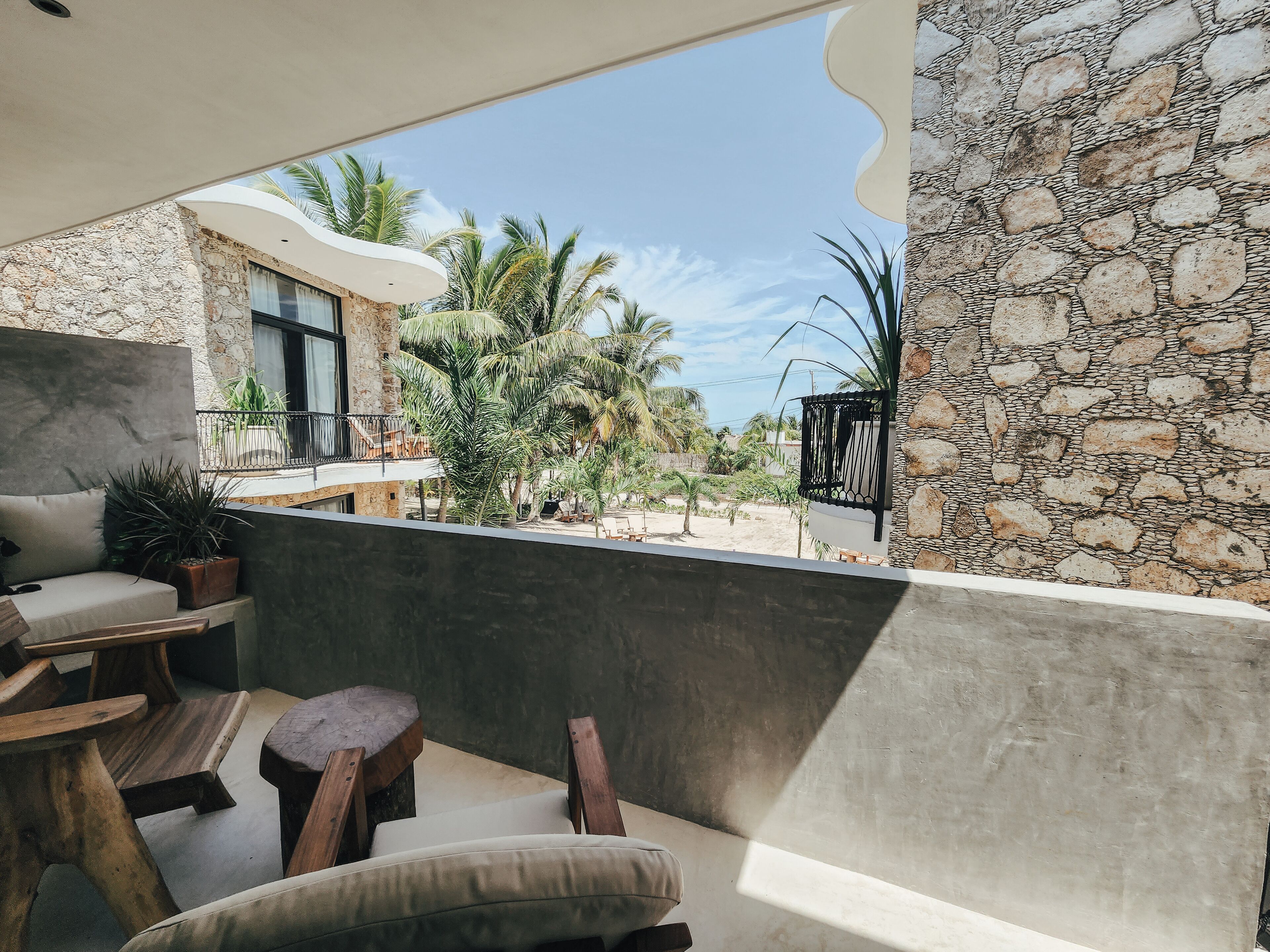 A balcony with a stone wall, a cushioned seating area, and a view of palm trees and a building.