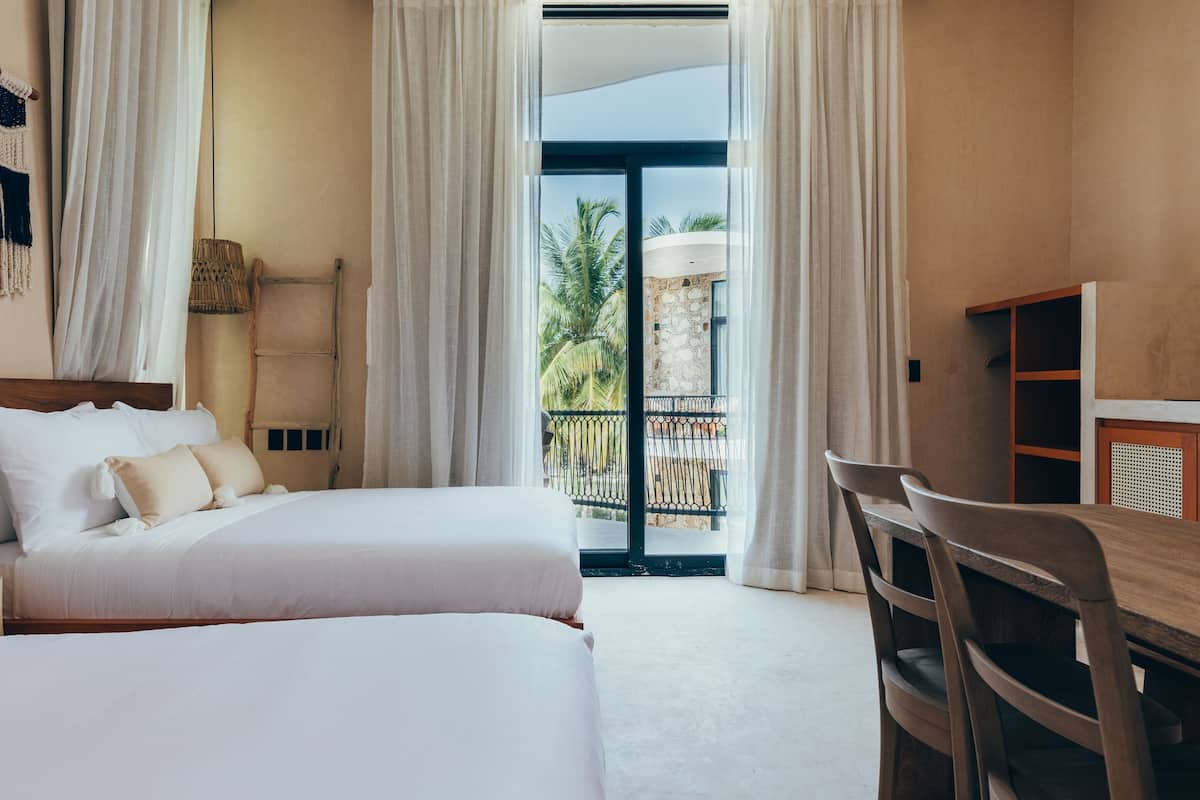 A hotel room with a bed, a desk, and a view of a balcony with palm trees.