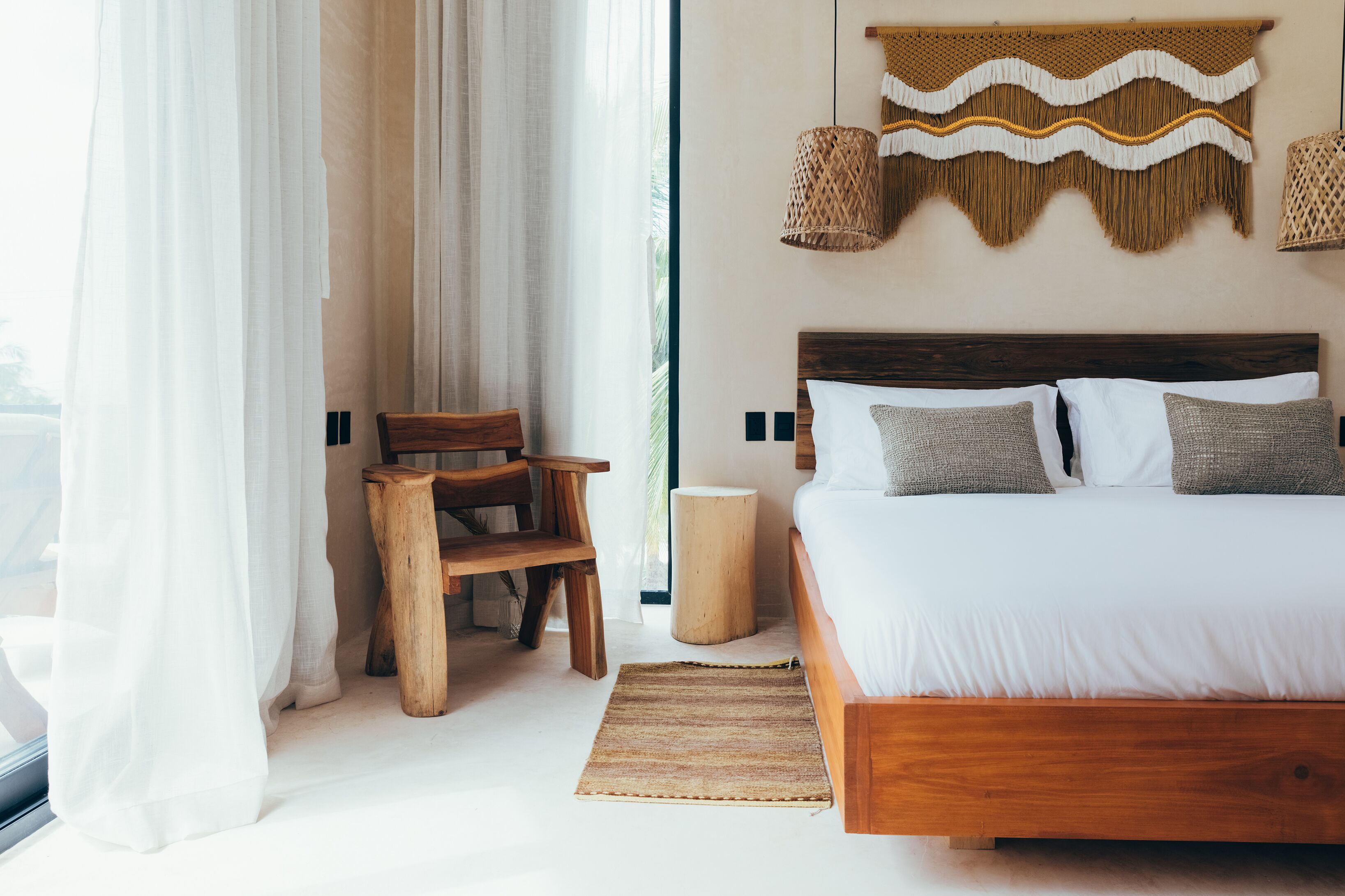 A bedroom with a wooden bed, white bedding, a woven wall hanging, and a small wooden stool.
