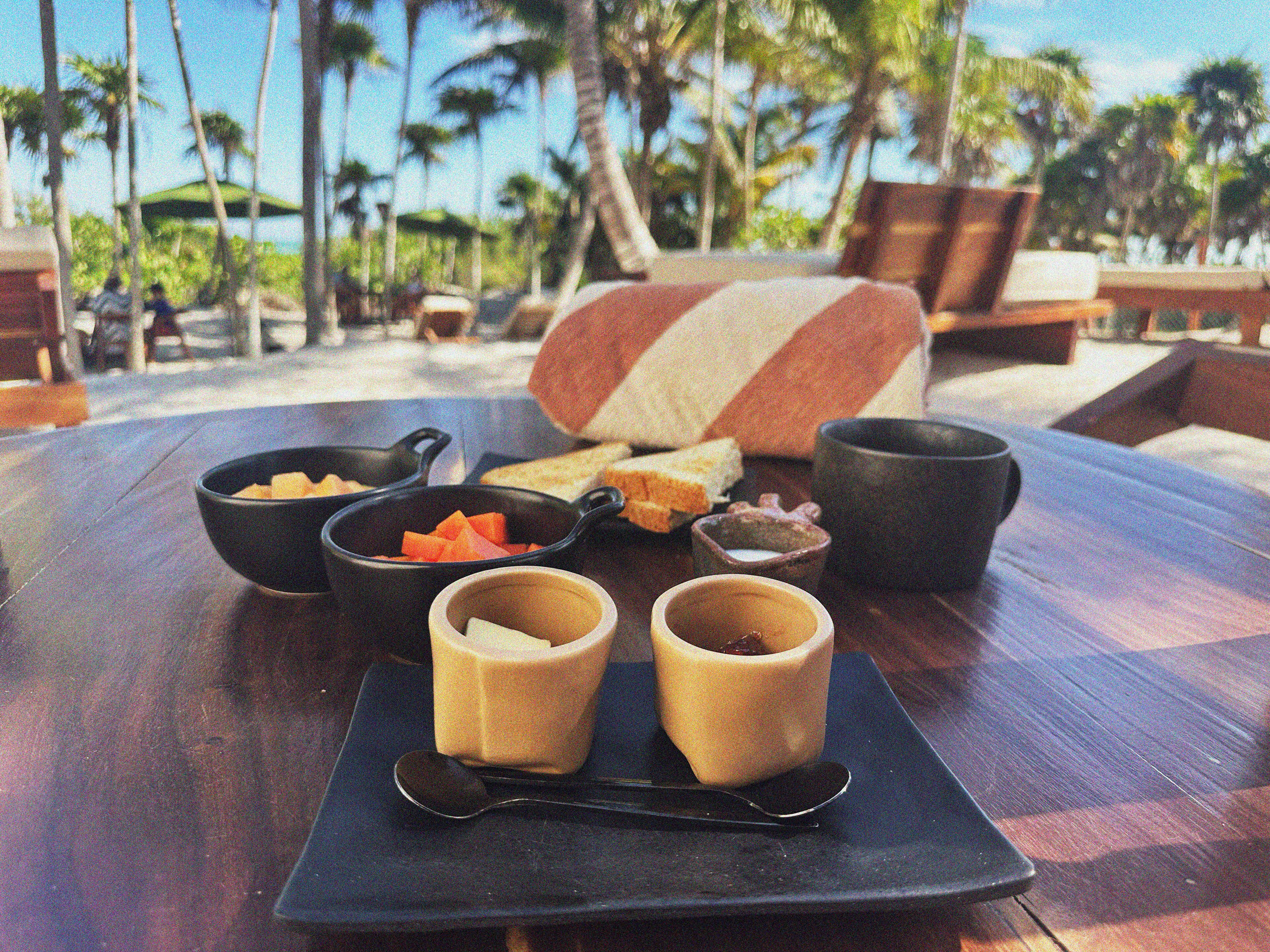 A table with a meal setup including bowls, a cup, and a spoon, set against a backdrop of a beach with palm trees and lounge chairs.