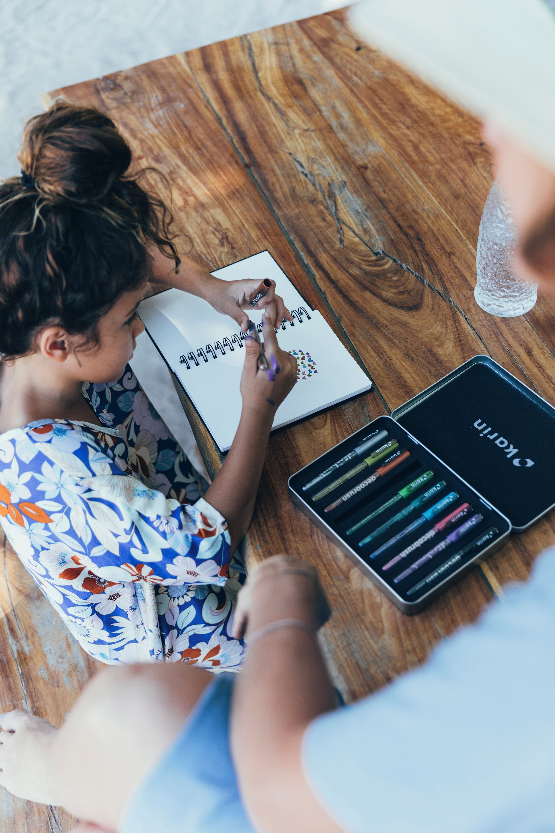 A person sitting at a wooden table with a notebook, a pen, and a box of colorful markers.