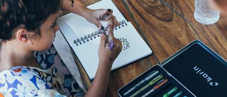 A person sitting at a wooden table with a notebook, a pen, and a box of colorful markers.