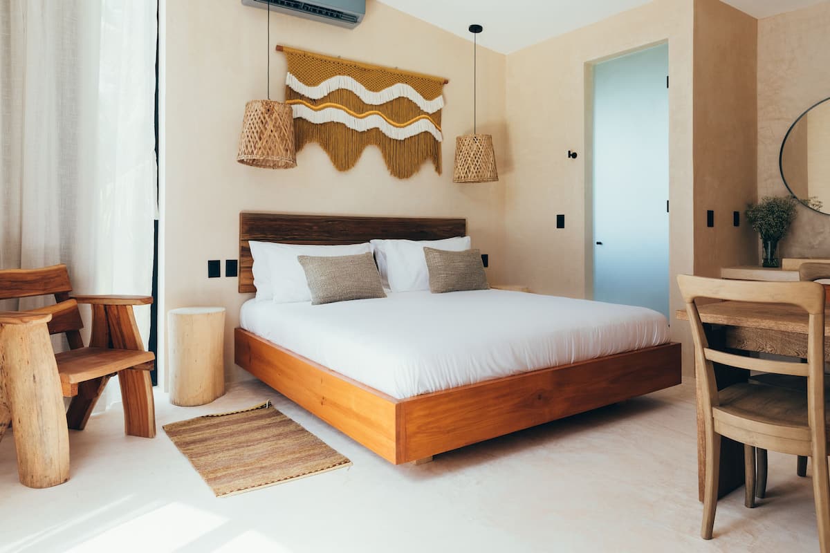 A bedroom with a wooden bed, two hanging lamps, a woven tapestry, a wooden chair, a round wooden table, and a small round stool.
