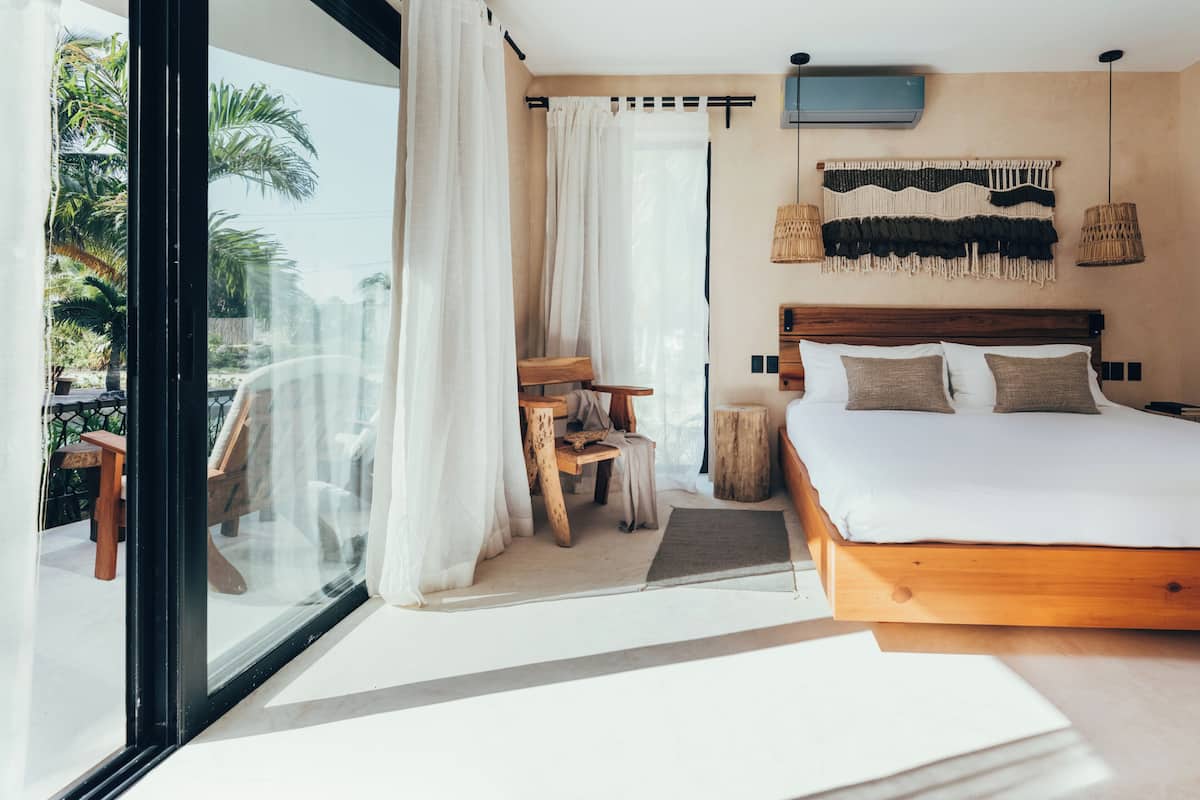 A bedroom with a large bed, a wooden headboard, a TV mounted on the wall, and a view of palm trees through the window.