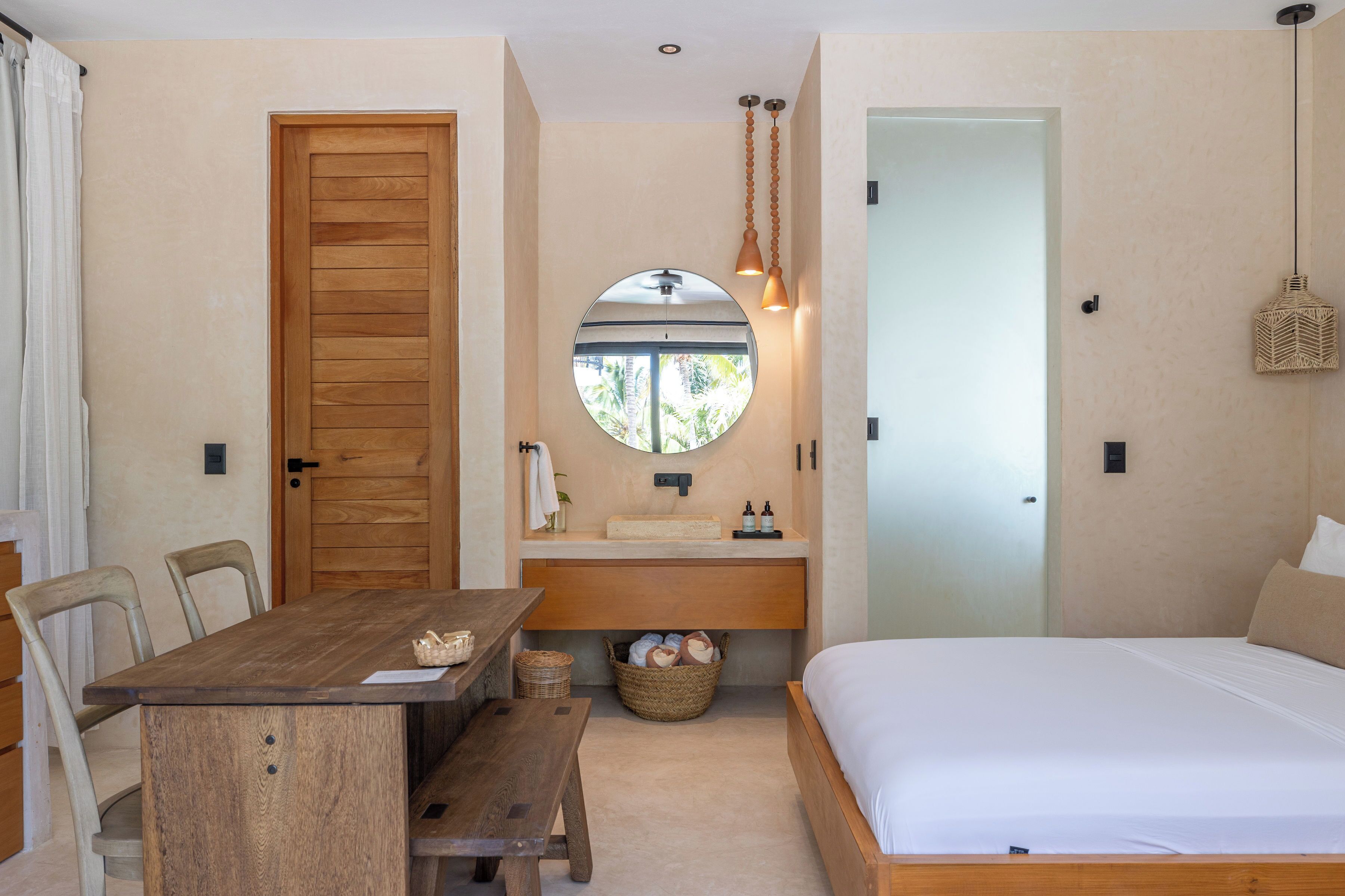 A bedroom with a wooden bed, a round mirror above a sink, a wooden dining table, and a wooden door.