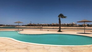 Una piscina de diseño curvo, rodeada por una terraza de piedra. Palmeras y sombrillas se ven al fondo.
