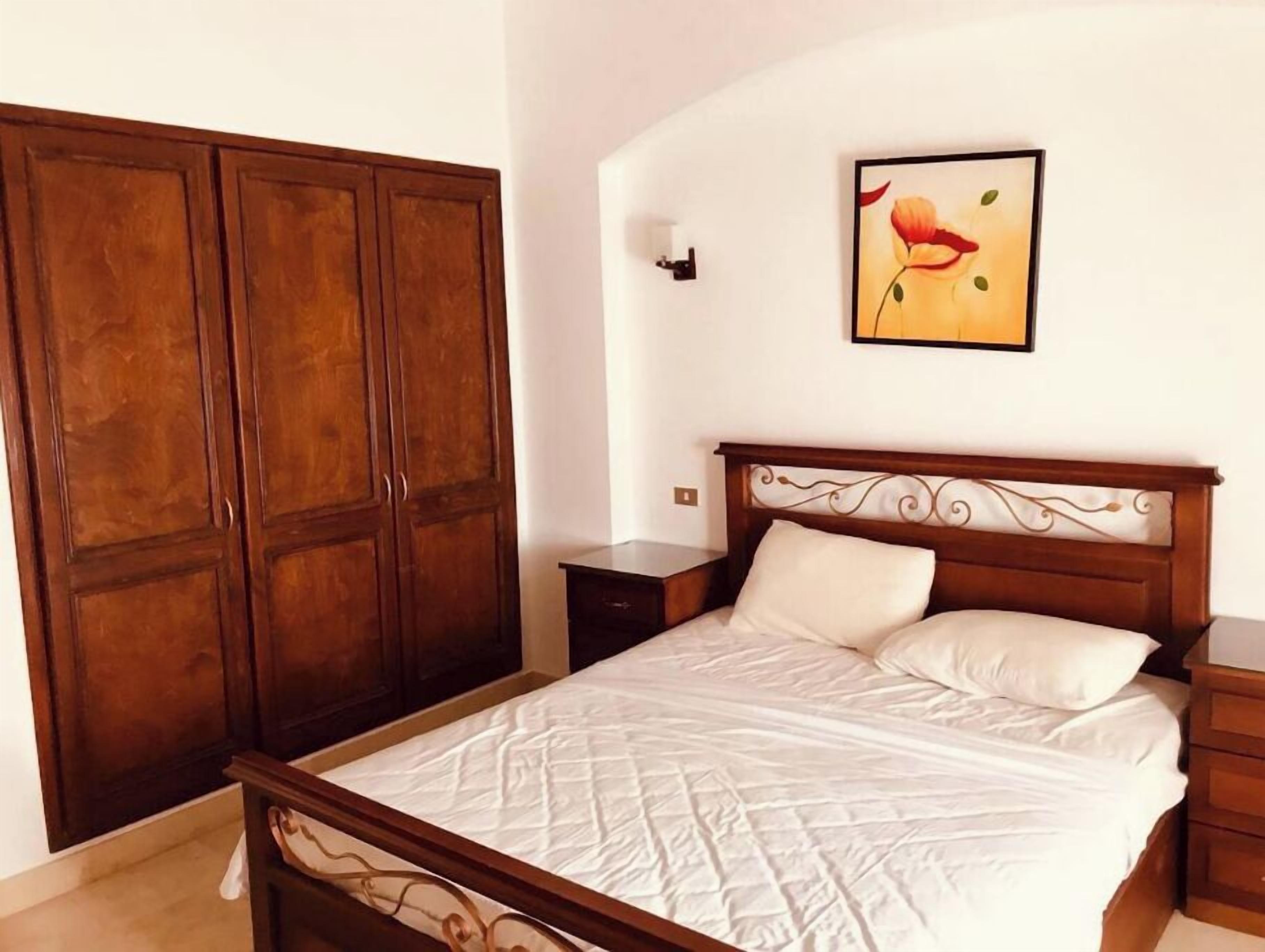 A bedroom with a wooden bed frame, a quilted mattress, a nightstand, a wooden wardrobe, and a framed picture on the wall.