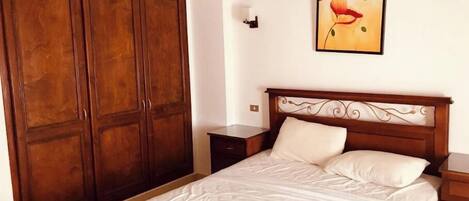 A bedroom with a wooden bed frame, a quilted mattress, a nightstand, a wooden wardrobe, and a framed picture on the wall.