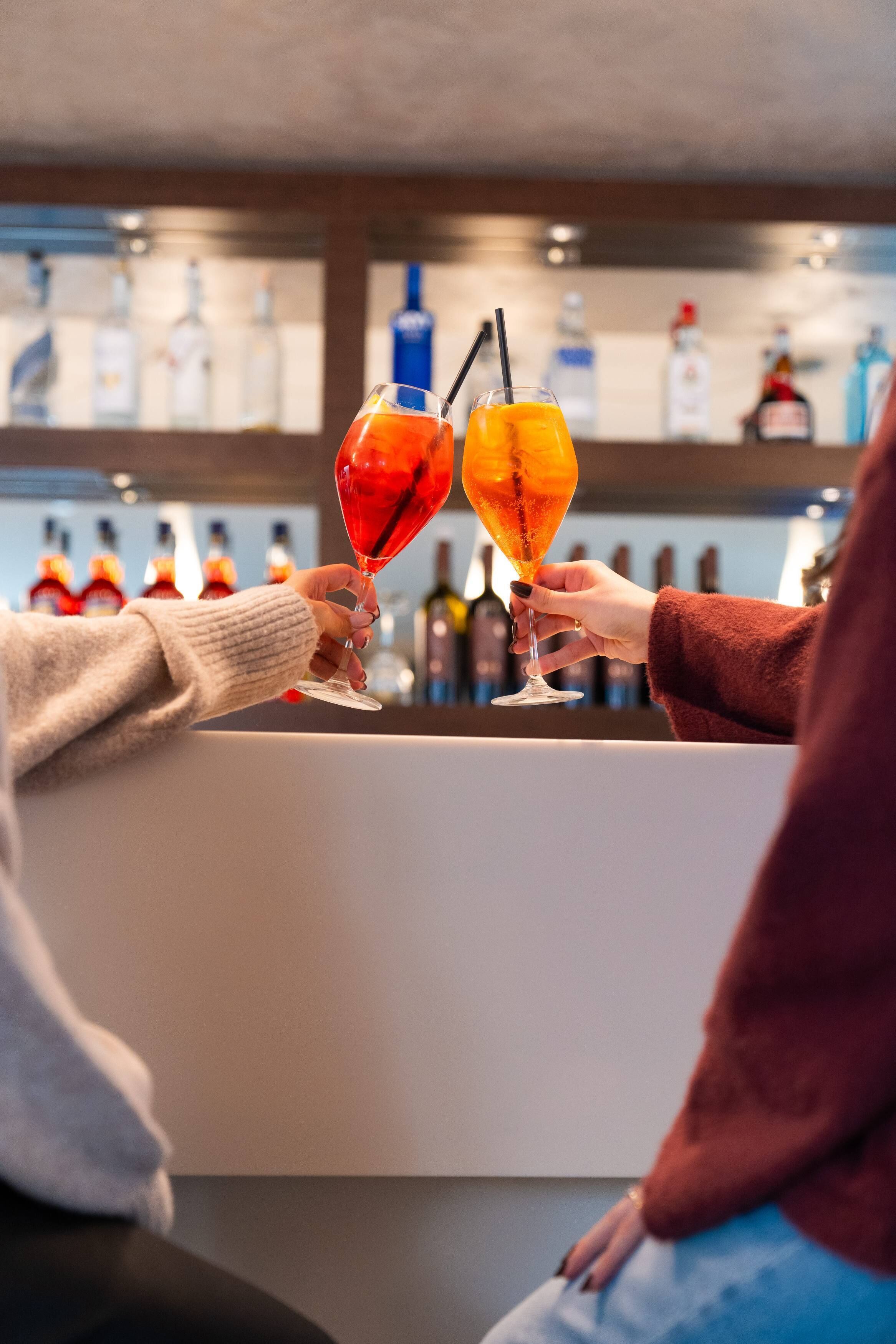 Two people holding cocktail glasses at a bar with a variety of bottles in the background.