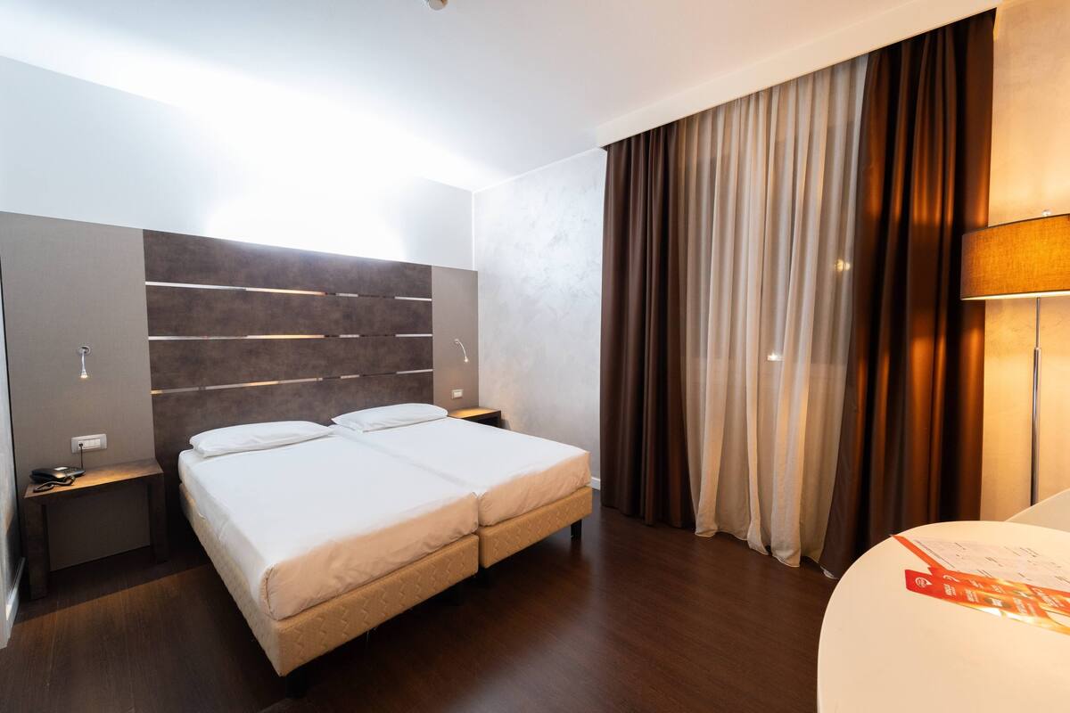 A hotel room with a large bed, a bedside table, a lamp, and a curtain.