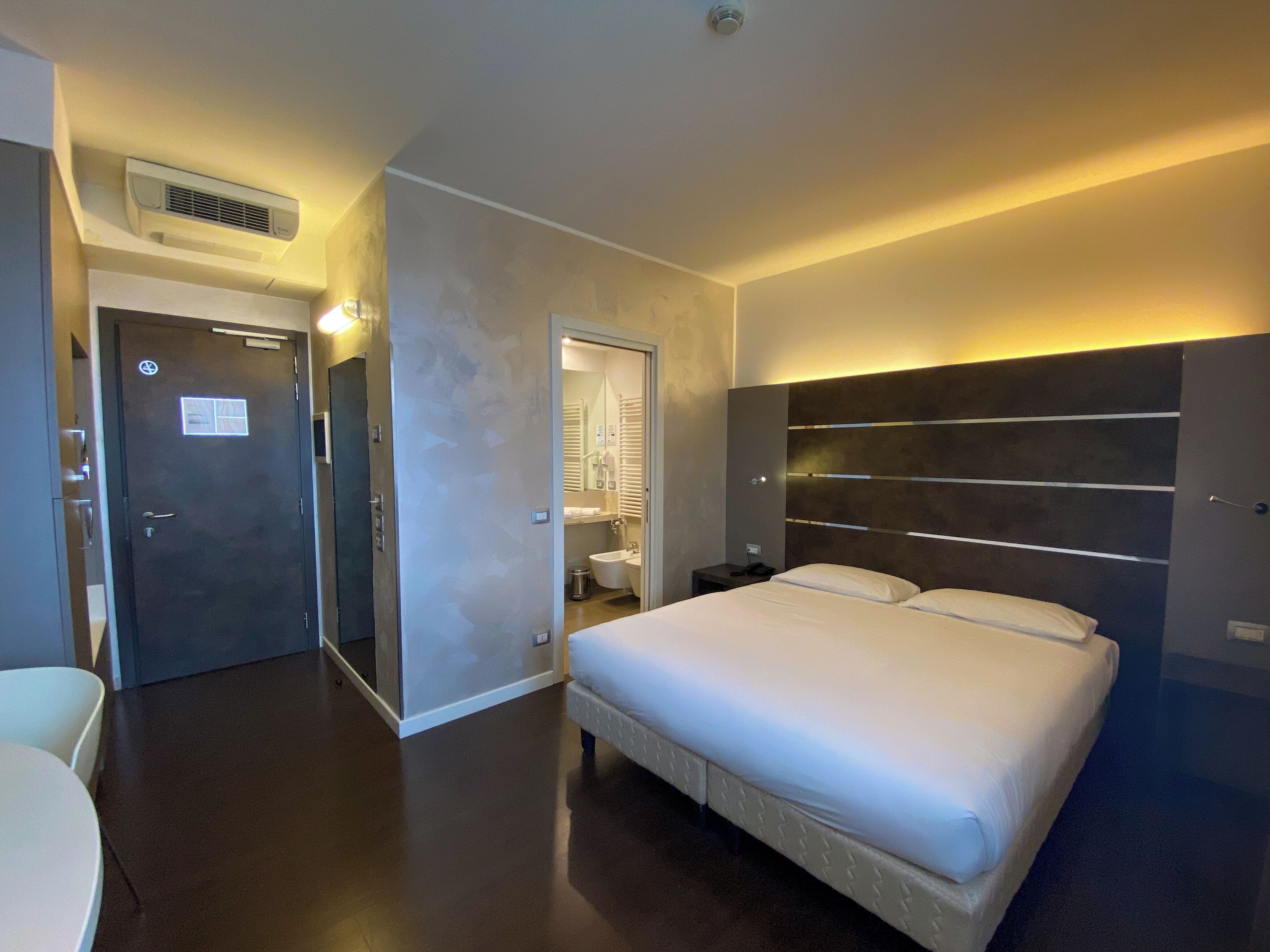 Una habitación de hotel moderna con una cama grande, una mesita de noche, un espejo y un equipo de aire acondicionado.
