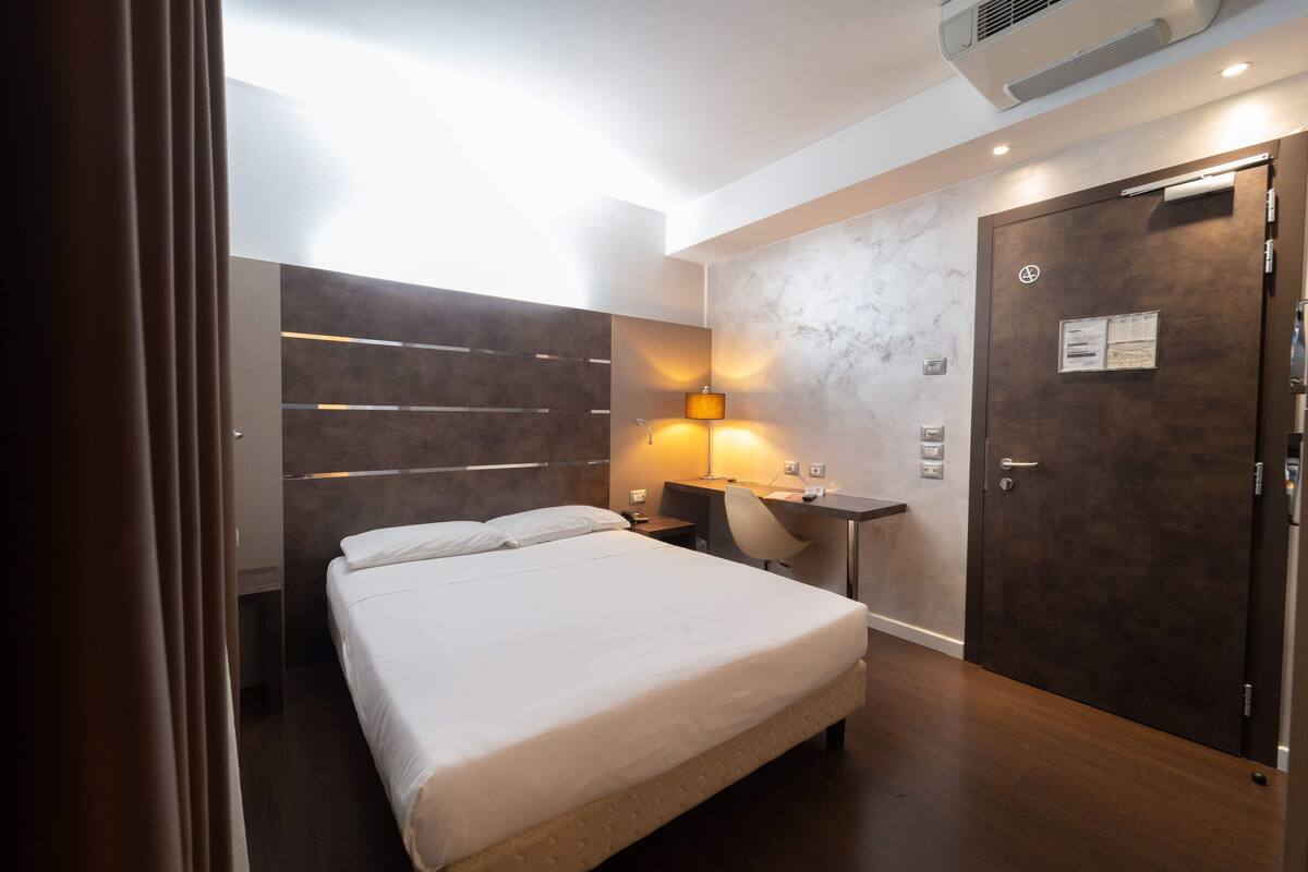 A hotel room with a bed, a desk, a chair, a wall-mounted lamp, and a door with a lock.