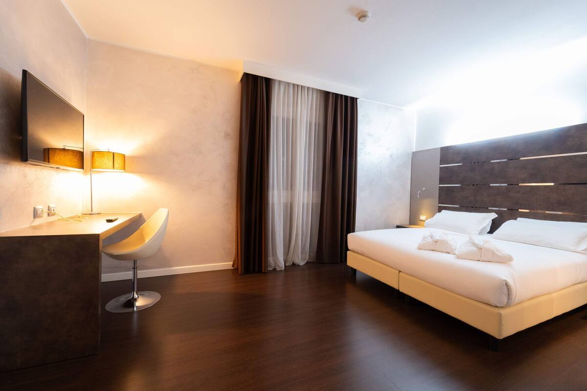 A modern hotel room with a large bed, a desk with a chair, a mirror, and a window with curtains.