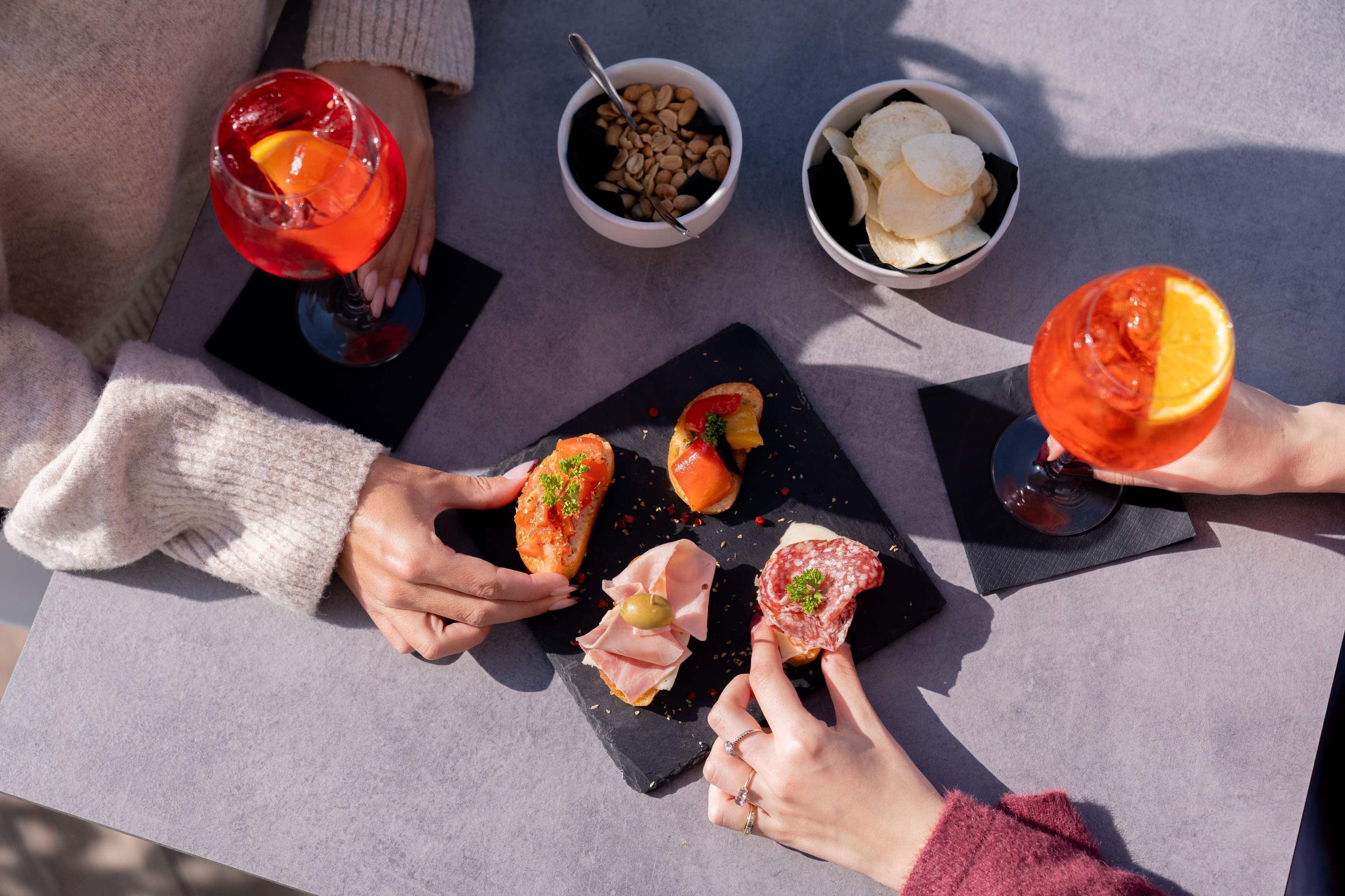 Ein Tisch mit Snacks und Getränken, darunter eine schwarze Schiefertafel mit verschiedenen Vorspeisen sowie zwei Gläser mit roten Getränken und Orangenscheiben.