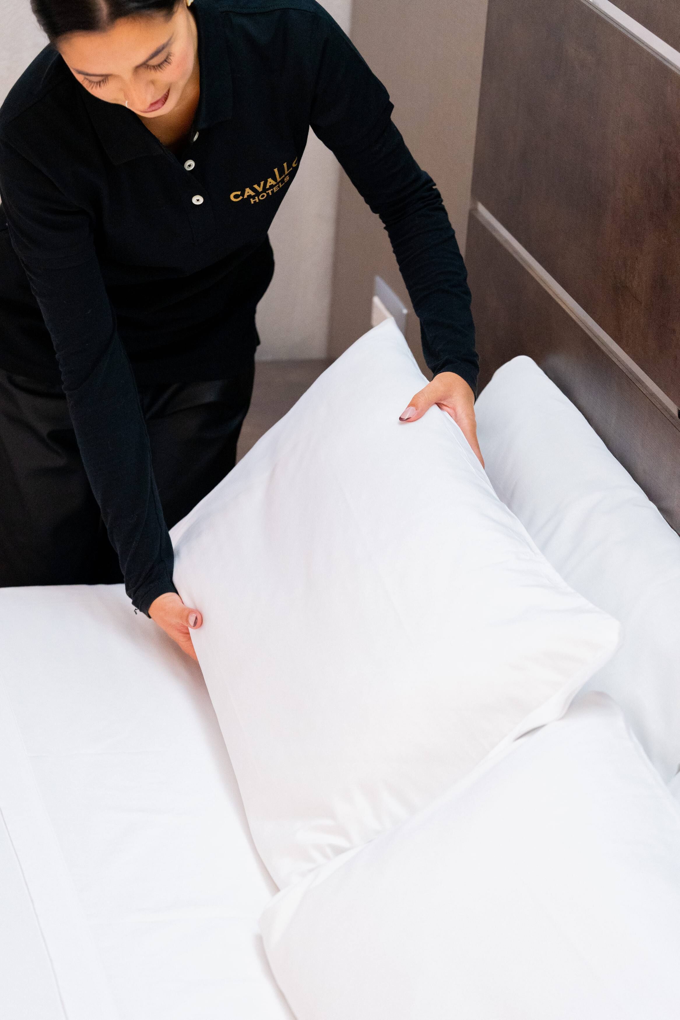 Una persona acomodando un almohadón blanco sobre una cama en una habitación de hotel.