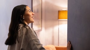Una persona abre una puerta en una habitación con una cama y una lámpara al fondo.