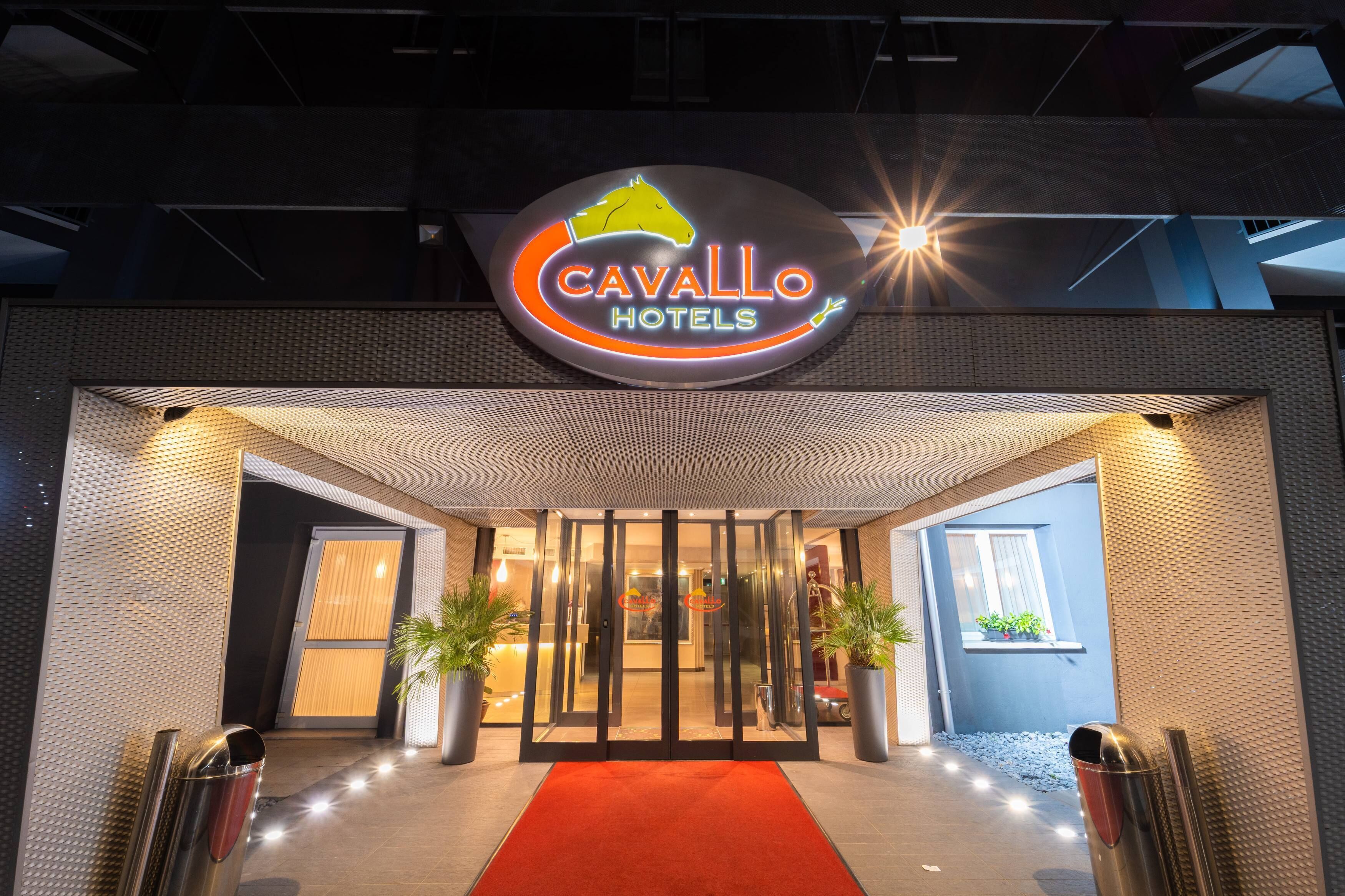 La entrada de Cavallo Hotels, con alfombra roja, puertas de cristal y plantas en macetas.