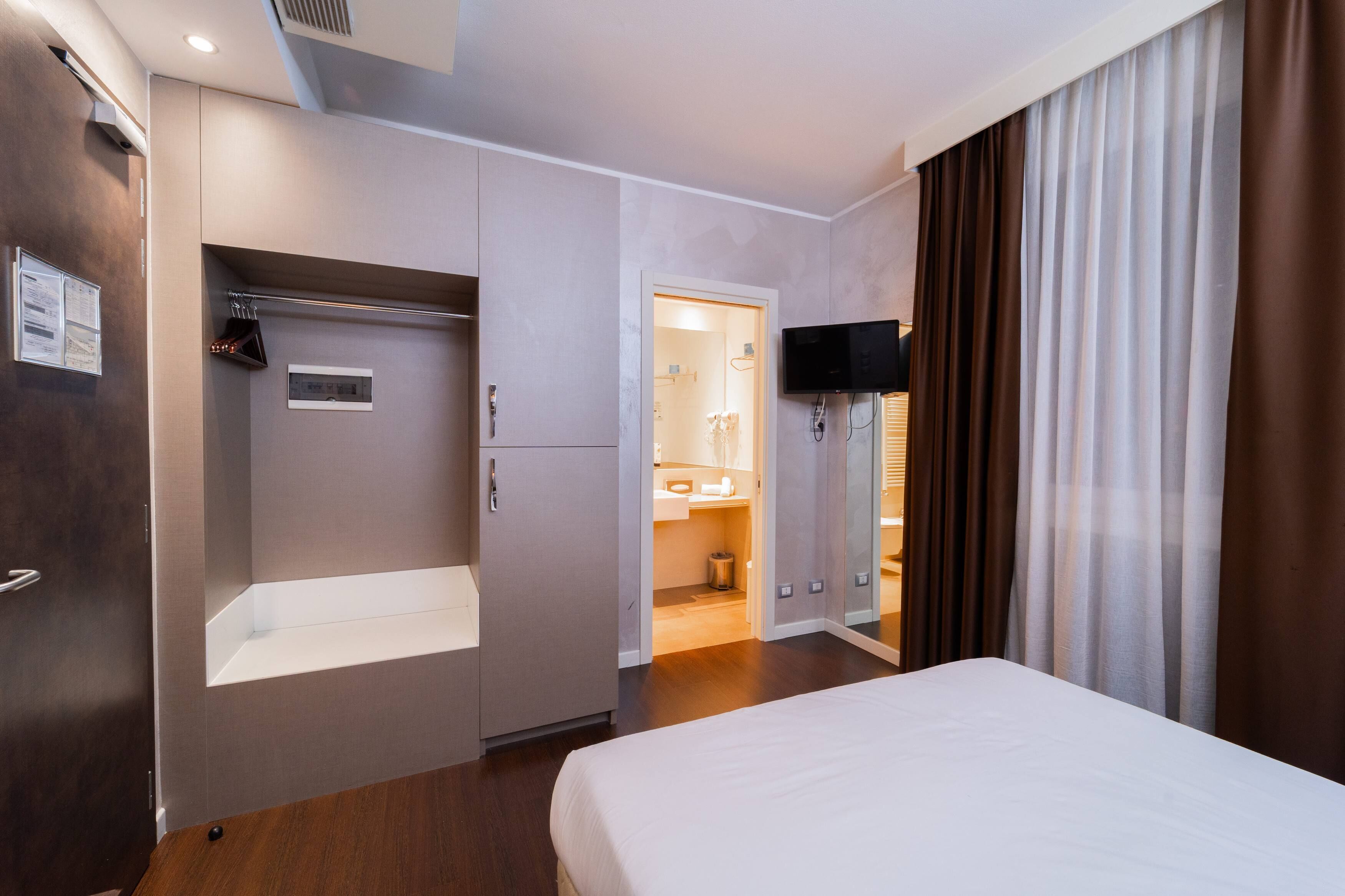 Habitación de hotel con cama, armario, televisión y baño visible a través de una puerta abierta.