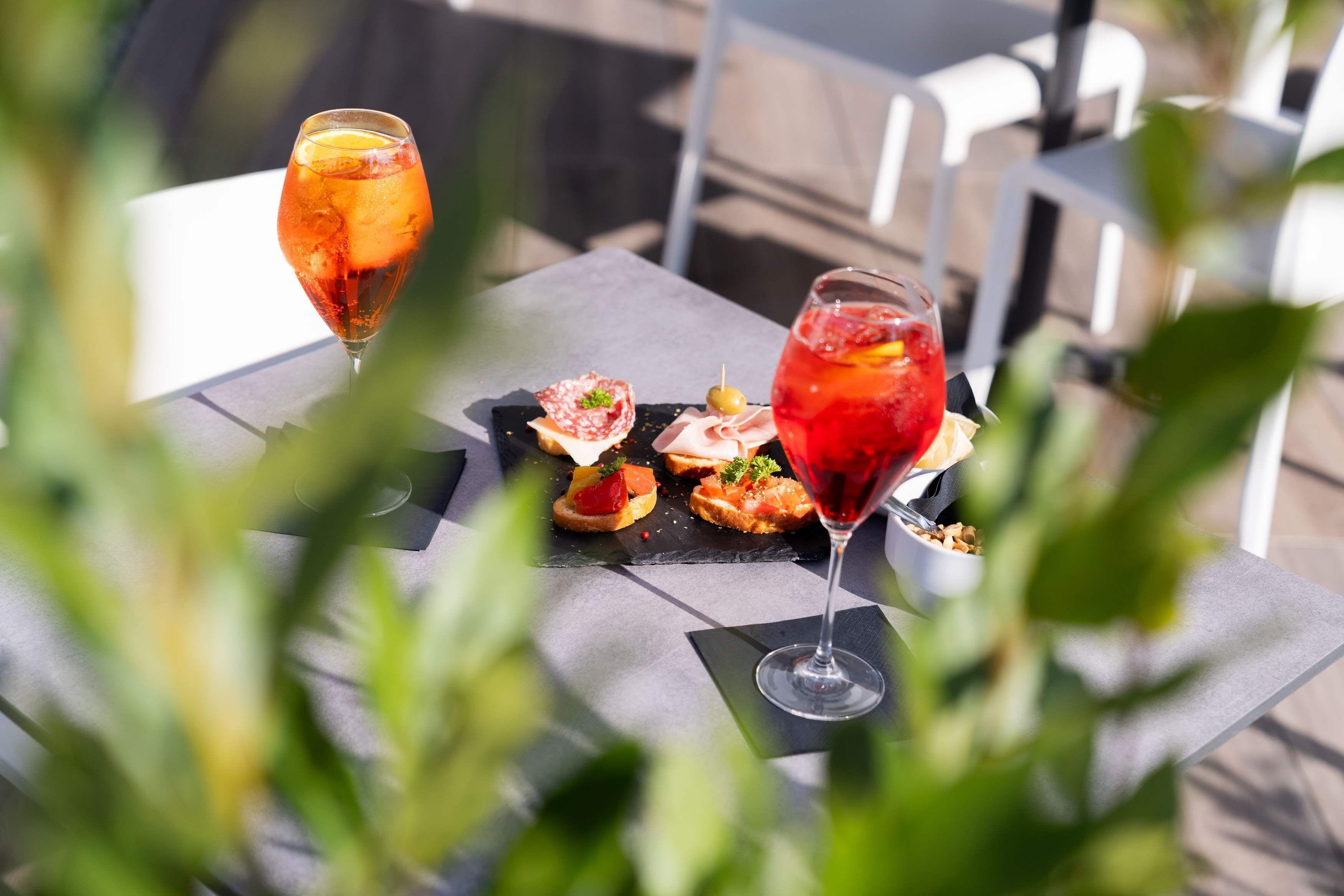A table with two glasses of red drink, a black slate board with food items, and a white chair in the background.