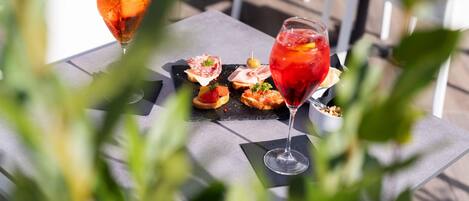 A table with two glasses of red drink, a black slate board with food items, and a white chair in the background.