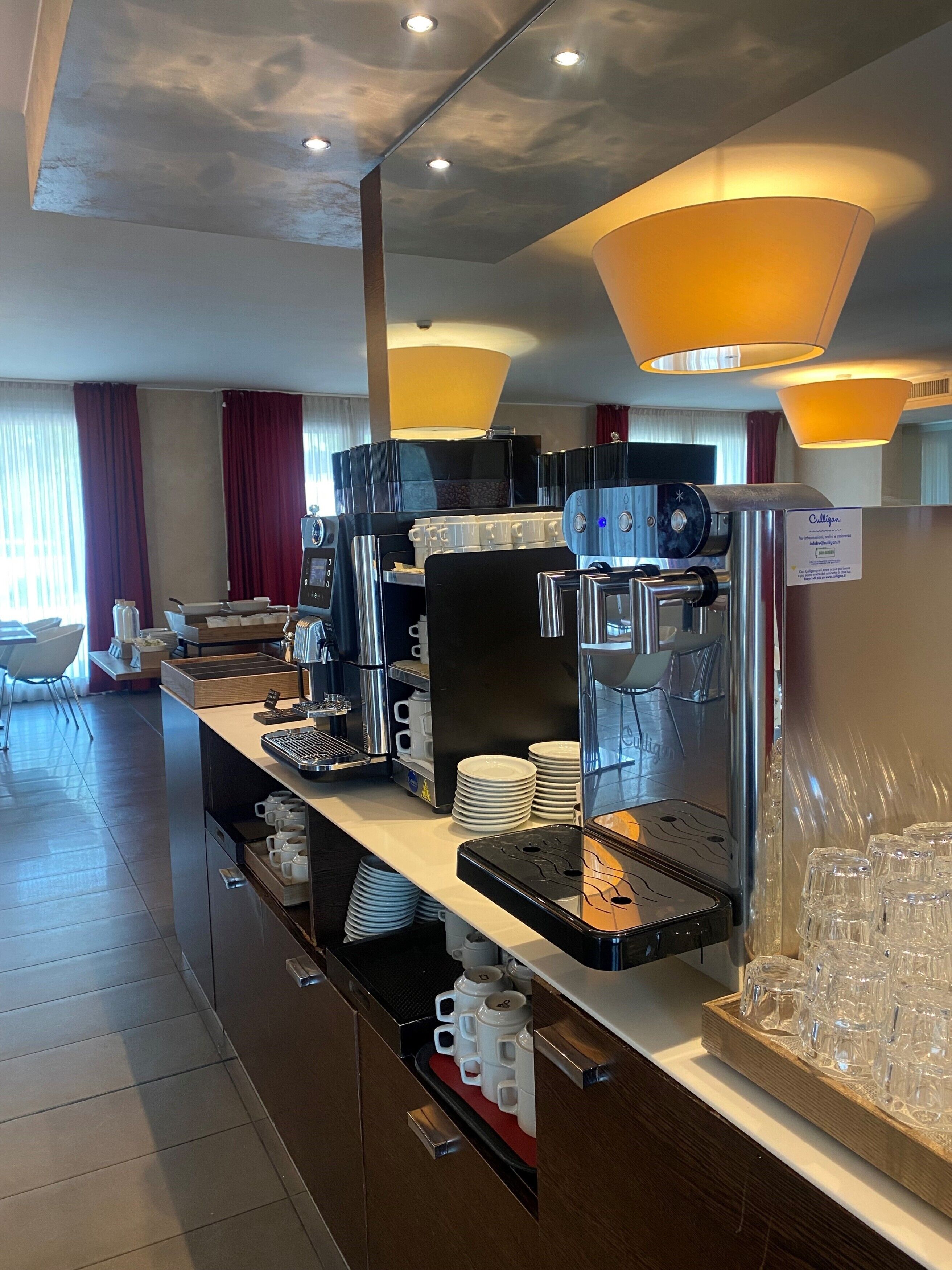A coffee station with multiple coffee machines, a stack of white plates, and a tray of clear glasses.